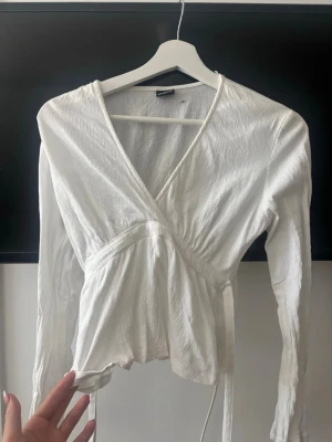 Vit omlottopp från Gina Tricot - Säljer en vit omlottopp från Gina Tricot i lätt och skönt material. Toppen har långa ärmar, v-ringning och knytband i midjan för en snygg passform. Perfekt att styla med jeans eller kjol för en trendig look.
