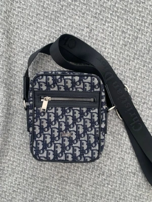 Mörkblå axelväska från Dior - Snygg axelväska från Dior i mörkblått och vitt med klassiskt monogrammönster. Väskan har en justerbar svart axelrem med Dior-logga, en dragkedjeficka framtill och silverfärgade detaljer. Perfekt för dig som vill ha en trendig och praktisk accessoar.