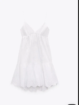 Vit somrig klänning med broderi - Söt vit kortklänning med tunna axelband och v-ringning. Klänningen har broderade detaljer och volangkant nertill, vilket ger en romantisk känsla. Perfekt för varma dagar och midsommar. Tillverkad i bomull för en luftig och bekväm känsla.