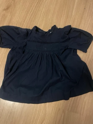Mörkblå blus med volang från Gina Tricot - Snygg mörkblå blus från Gina Tricot med volangdetaljer på axlarna och smockad överdel. Blusen har korta puffärmar och rund halsringning. Tillverkad i ett mjukt, tunt tyg som ger en luftig känsla. Perfekt för en trendig och avslappnad look.