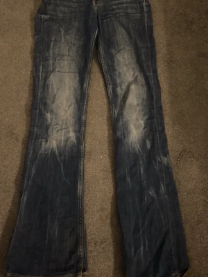Blå bootcut jeans med slitningar - Säljer ett par blå bootcut jeans med tydliga slitningar och ljusa tvättade partier framtill och baktill. Jeansen har klassisk femficksdesign och råa kanter vid bensluten. Perfekta för dig som gillar en avslappnad och vintageinspirerad stil.