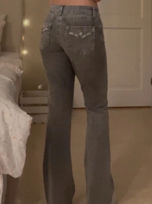 Grå bootcut jeans med fickdetaljer - Säljer ett par grå bootcut jeans med snygga fickdetaljer bak och knappar. Jeansen har normal passform upptill och utsvängda ben. Liten missfärgning nedtill på ena benet, syns på sista bilden💗