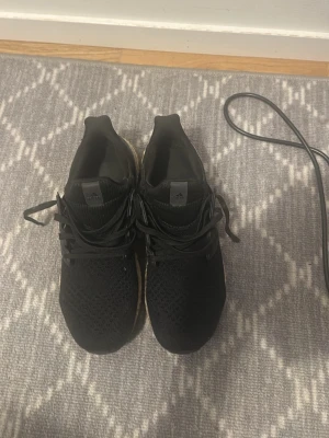 Svarta Adidas sneakers i mesh - Säljer ett par svarta Adidas sneakers med stickad mesh-överdel och snörning. Skorna har en vit sula med svart ovandel och klassisk Adidas-logga på plösen. Perfekta för dig som gillar sportig och stilren look.