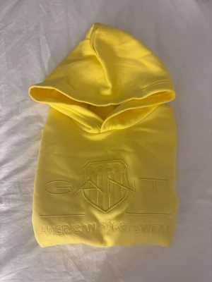 Gul hoodie från GANT - Snygg gul hoodie från GANT med broderad logga och texten 'American Sportswear' på bröstet. Klassisk huva och ribbade muddar vid ärmslut och nederkant. Hoodien är i storlek 158/164 och har aldrig blivit använd men saknar prislapp💕