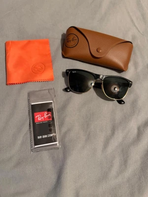 Ray-Ban Clubmaster svarta solglasögon - Snygga Ray-Ban Clubmaster solglasögon i svart med gulddetaljer. Klassisk retroform med mörka glas. Kommer med originalfodral, putsduk och Ray-Ban certifikat. Perfekta för att lyfta din stil i solen!