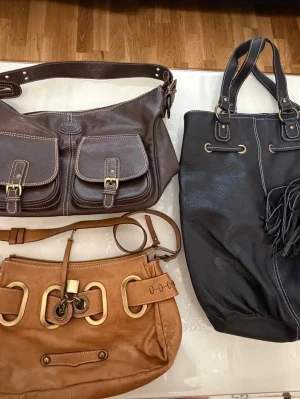 Tre handväskor i läderlook, Från Tod’s , Antoni och Rizoo  - Säljer tre snygga handväskor: en mörkbrun med dubbla fickor och spännen, en svart med fransdetalj och långa handtag samt en ljusbrun med stora guldfärgade ringar och axelrem. Alla i läderimitation och perfekta för dig som gillar klassiska men trendiga accessoarer. Kan sälja separat och tillsammans 