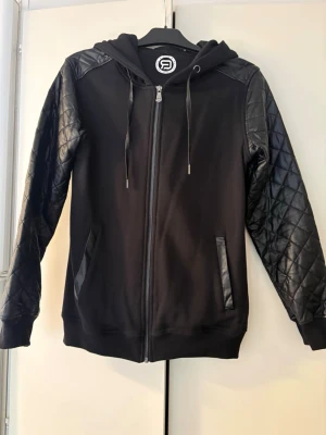 Svart hoodie med quiltade ärmar - Säljer en svart hoodie med dragkedja och huva. Ärmarna är quiltade i ett glansigt material som ger en schysst kontrast till resten av hoodien. Fickor med detaljer i samma material som ärmarna. Perfekt för dig som gillar streetwear vibes.