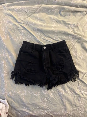 Svarta fransiga jeansshorts - Svarta jeansshorts med hög midja och slitna detaljer framtill. Shortsen har fransig kant nedtill och klassisk femficksdesign. Perfekta för en avslappnad och trendig look.