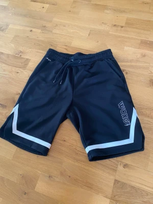 Svarta träningsshorts med snörning - Snygga svarta träningsshorts med vita detaljer och snörning i midjan. Shortsen har elastisk midja, fickor på sidorna och ett tryckt märke på ena benet. Perfekta för sport eller chill dagar.
