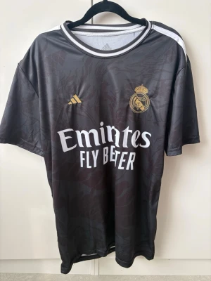 Real Madrid Ronaldo matchtröja svart - Snygg svart Real Madrid matchtröja från Adidas med guldiga detaljer och klubbmärke på bröstet. Tryck med 'Emirates Fly Better' fram och 'Ronaldo 7' bak. Tillverkad i lätt och ventilerande material, perfekt för fotboll eller träning.