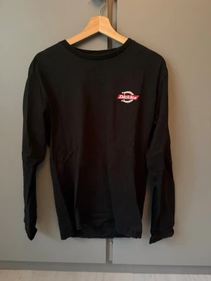 Svart långärmad tröja Dickies - Svart långärmad tröja från Dickies med rund hals och tryckt logga i vitt och rött på både bröstet och ryggen. Tillverkad i mjukt bomullsmaterial och har en avslappnad passform. Perfekt för en streetwear-look.