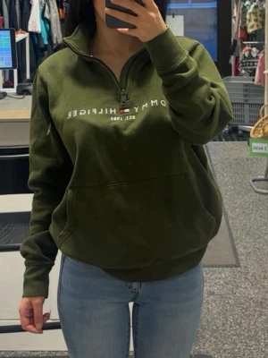 Olivgrön Tommy Hilfiger hoodie - Snygg olivgrön hoodie från Tommy Hilfiger med halv dragkedja och stor magficka. Tryckt logga och flagga på bröstet. Mjuk och skön bomullsblandning, perfekt för en avslappnad stil. Ribbstickade muddar vid ärmar och nederkant.