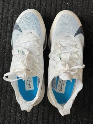 Vita och blå Nike sneakers - Snygga vita sneakers från Nike med blå och grå detaljer. Ovandelen är i mesh för bra andningsförmåga och sulan har en synlig luftdämpning. Skorna har klassisk snörning och en sportig look som passar perfekt till streetwear.