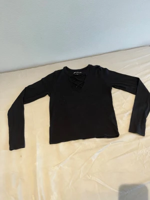 Svart långärmad topp med snörning - Snygg svart långärmad topp med snörning framtill i halsringningen. Modellen är croppad och har en slim passform som passar perfekt till både jeans och kjol. Enkel men trendig design som funkar till många olika stilar.