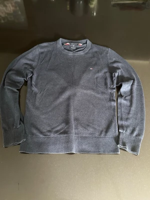 Mörkblå stickad tröja Tommy Hilfiger - Mörkblå stickad tröja från Tommy Hilfiger i mjuk bomullsblandning med kashmir. Klassisk rund halsringning, ribbade muddar och liten broderad logga på bröstet. Perfekt för dig som gillar stilrena och bekväma plagg.