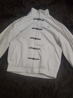 Vit Adidas jacka med snörning - Jätte snygg Adidas jacka i cremevit, storlek M i kinesiskt nyårsstil med dragkedja och dekorativa spännen. Har använts ett fåtal gånger, men väl omhändertagen! Mått: Längd: 72,5 cm Armhålsvidd: 57 cm Ytterärmlängd: 55cm För fler frågor, kontakta gärna mig! 🌻