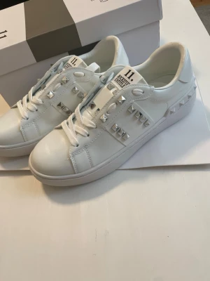 Valentino dam skor  - Snygga vita sneakers med silverfärgade pyramidnitar och vita skosnören. Skorna har en stilren design med dekorativa detaljer på sidorna och bakkappan. Ovandelen är i slätt material och sulan är vit. Perfekta för dig som gillar en edgy och trendig look.