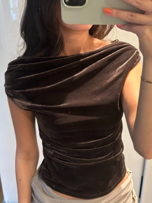 Brun sammets topp med drapering - Snygg brun topp i mjuk sammet med draperad design och asymmetrisk halsringning. Toppen är ärmlös och har en tight passform som framhäver siluetten. Perfekt för dig som vill ha en unik och trendig look. Storlek S men är lite tight så passar absolut storlek xs också