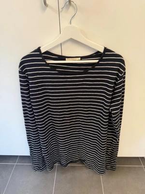 Randig långärmad t-shirt Nudie Jeans, stl M - Säljer en svart långärmad t-shirt från Nudie Jeans med vita horisontella ränder. Toppen har en klassisk rund halsringning och är gjord i mjuk bomull som känns skön mot huden. Perfekt för en avslappnad och stilren look.