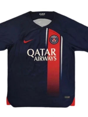 PSG matchtröja blå/röd Nike - Snygg PSG matchtröja i mörkblått med röd vertikal rand och vita detaljer. Tröjan har klubbmärke och Qatar Airways-logga på bröstet samt Nike-logga i rött. Tillverkad i lätt och ventilerande material, perfekt för fotboll eller träning.