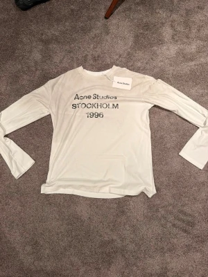Vit långärmad topp Acne Studios - Vit långärmad topp från Acne Studios med tryck 'STOCKHOLM 1996' på bröstet. Tunn och mjuk kvalitet med rund halsringning. Perfekt för en clean och stilren look.