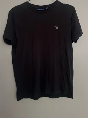 Svart t-shirt från Gant - Svart t-shirt från Gant med liten logga och sköld på bröstet. Klassisk rund halsringning och korta ärmar. Enkel och stilren design som passar till det mesta. Tillverkad i mjuk bomull för en bekväm känsla.