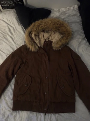 Brun bomberjacka  - Snygg brun bomberjacka med stor luva kantad av beige fuskpäls. Jackan har ribbade muddar, två framfickor med knappar och quiltat innerfoder. Perfekt för kalla dagar och ger en cool streetwear-look.