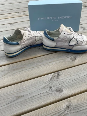Vita Philippe Model sneakers med blå detaljer - Snygga vita sneakers från Philippe Model med blå detaljer på häl och sula. Skorna har en klassisk stil  samt vita skosnören. Perfekta för dig som gillar stilrena och sportiga sneakers. Boxen ingår