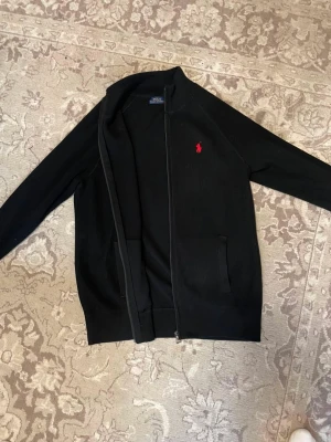 Svart ziptröja från polo - Snygg svart ziptröja från Ralph Lauren med röd broderad logga på bröstet. Tröjan har hög krage, två fickor framtill och är tillverkad i ett mjukt, stickat material. Perfekt för en stilren och avslappnad look.