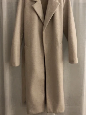Beige lång kappa - Stilren beige kappa med klassisk krage och långa ärmar. Kappan har rak passform och dolda fickor i sidorna. Perfekt för en trendig och minimalistisk look under kyliga dagar.