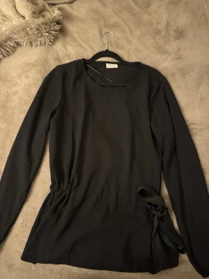 Svart långärmad topp med knyt - Snygg svart långärmad topp med rund halsringning och knyt i sidan som ger en extra detalj. Perfekt till jeans eller kjol för en clean och stilren look. Materialet känns mjukt och följsamt.
