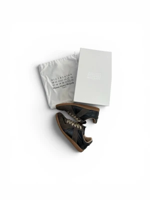 Maison Margiela svarta sneakers - Snygga sneakers från Maison Margiela i svart läder och mocka med beige snörning och gummisula. Klassisk låg modell med stilren design och diskreta detaljer. Perfekta för dig som gillar minimalistisk och trendig stil.