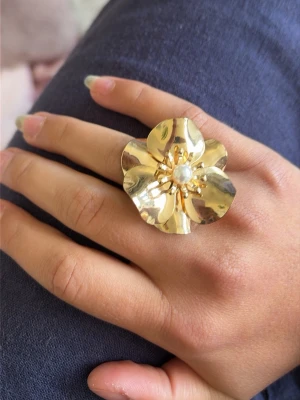 Guldfärgad blomring med pärldetalj - Säljer en stor och iögonfallande ring i guldfärg formad som en blomma. Ringen har fem blad och en vit pärla i mitten som ger en elegant touch. Perfekt för dig som vill sticka ut med dina accessoarer.