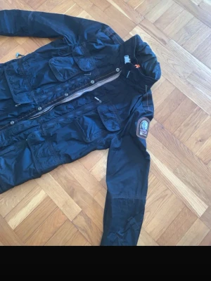 Svart vindjacka från Parajumpers - Snygg svart vindjacka från Parajumpers med flera fickor, hög krage och dragkedja. Jackan har patch på ärmen och orange detalj i nacken. Tillverkad i lätt, glansigt material som står emot vind. Perfekt för dig som vill ha en sportig och funktionell look.