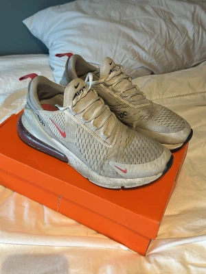 Nike Air Max 270 grå sneakers - Nike Air Max 270 sneakers i grått med röda detaljer och stor synlig Air-enhet i hälen. Ovandelen är i mesh för bra ventilation och har klassisk snörning. Swoosh-logga på sidan och tå, samt AIR270-tryck på plösen. Perfekta för en sportig och trendig look.