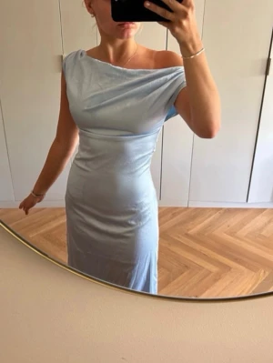 Ljusblå oneshoulder långklänning - Säljer en elegant ljusblå långklänning med oneshoulder-design. Klänningen har en smickrande, figurnära passform och är tillverkad i ett glansigt, mjukt material som ger en lyxig känsla. Perfekt för dig som vill sticka ut med en stilren look. Några små fläckar från vatten. Borde försvinna med rätt tvätt. Använd en gång. Är 170 cm lång och brukar ha kläder i kring storlek S.