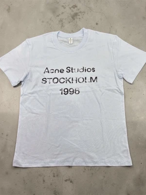 Vit Acne Studios t-shirt Stockholm - Ljusvit t-shirt från Acne Studios med tryck 'Acne Studios STOCKHOLM 1996' i svart framtill. Klassisk rund hals och korta ärmar. Perfekt för en clean och stilren look.