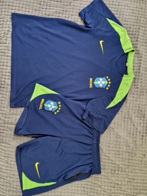 Blått Brasilien fotbollsställ Nike - Säljer ett blått Brasilien fotbollsställ från Nike med limegröna detaljer. Setet består av en t-shirt och shorts, båda med CBF-logga och Nike swoosh i gult. Perfekt för träning eller match. Materialet är lätt och andas, vilket gör det skönt att bära under aktivitet.