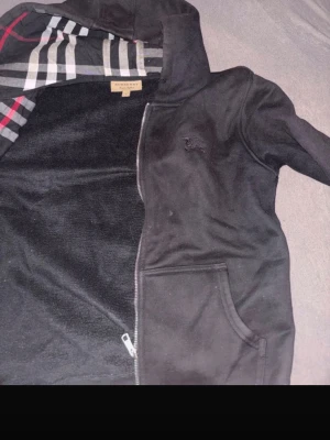 Svart hoodie med dragkedja Burberry - Snygg svart hoodie från Burberry med dragkedja och klassiskt rutigt foder i huvan. Hoodien har fickor framtill och en diskret broderad logga på bröstet. Materialet ser mjukt och bekvämt ut, perfekt för en chill och stilren look.