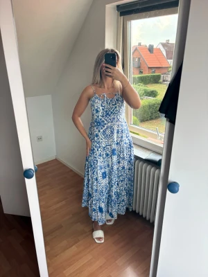 Blåvit långklänning med mönster - Säljer en blåvit långklänning med tunna axelband och knyt fram. Klänningen har ett vackert blommigt och ornamenterat mönster, luftig passform och är perfekt för varma dagar. Tillverkad i ett lätt material som känns skönt mot huden. 100% bomull!!