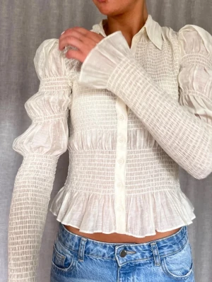 Vit smockad blus från & other stories  - Supersöt vit blus med smockdetaljer, puffiga långärmar och volangkant nedtill. Blusen har knappar framtill och är gjord i ett tunt, lätt material😊