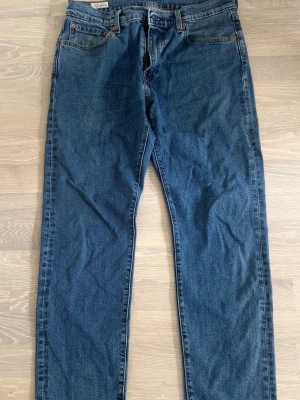 Blå raka jeans från Levi's - Klassiska blå jeans från Levi's 502 med rak passform och fem fickor. Jeansen har en djupblå tvätt och detaljer i kopparfärgade nitar. Tillverkade i slitstark denim med Levi's ikoniska bakfickor och patch bak i midjan. Storlek 33/32. 