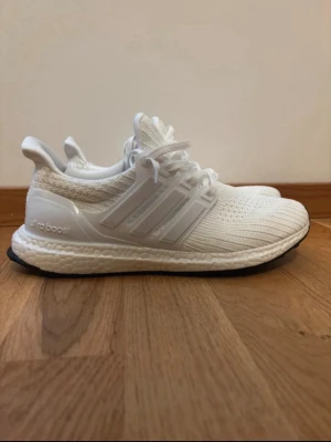 Vita Adidas Ultraboost sneakers - Säljer ett par helvita Adidas Ultraboost sneakers med stickad ovandel och klassiska tre ränder på sidan. Skorna har en tjock, responsiv Boost-sula och svart yttersula. Perfekta för dig som gillar stilrena och bekväma sneakers.