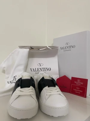 Valentino Garavani vita sneakers - Snygga vita sneakers från Valentino Garavani med svart detalj över snörningen. Tillverkade i läder och har klassisk låg siluett. Perfekta för dig som vill ha en clean och exklusiv look. Levereras med originalkartong och dustbag.