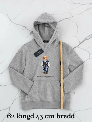 Grå hoodie Polo Bear Ralph Lauren - Grå hoodie från Ralph Lauren med Polo Bear-tryck framtill. Klassisk huva och känguruficka. Mjuk bomullsblandning och ribbade muddar. Perfekt för en avslappnad och trendig stil.