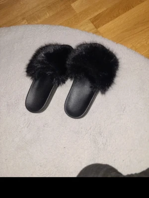 Sandaler - Säljer ett par svarta tofflor med bred rem täckt av mjuk fuskpäls. Sulan är i svart syntetmaterial och designen är öppen för en chill och trendig look. Perfekta för att glida runt hemma med extra stil. Aldrig använda 