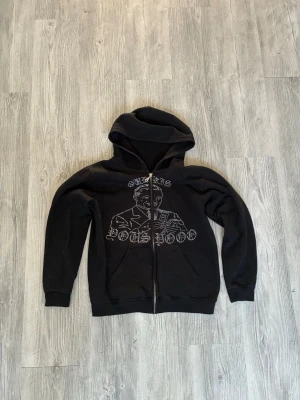 Svart hoodie med grafiskt tryck - Svart hoodie med dragkedja och stor huva. På framsidan finns ett grafiskt tryck av en person och texten 'CHRIS BOSS POOO'. Hoodien har två fickor framtill och ribbade muddar vid ärmar och nederkant. Perfekt för en chill och avslappnad stil.