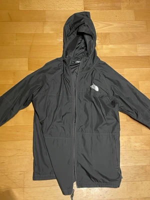 Svart vindjacka från The North Face - Snygg svart vindjacka från The North Face med huva och dragkedja framtill. Jackan har en minimalistisk design med logga på bröstet och är tillverkad i lätt, vindtätt material. Perfekt för blåsiga dagar och enkel att packa ner i väskan.