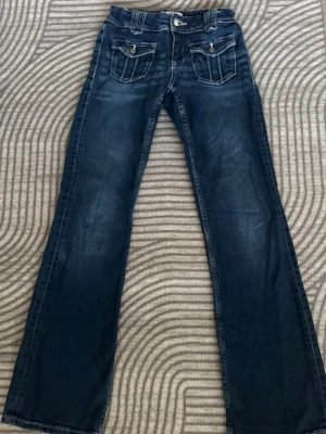 Mörkblå bootcut jeans med fickdetaljer - Pris går att diskuteras. Säljer ett par mörkblå jeans med bootcut ben och snygga fickor framtill med knappdetaljer. Klassisk femficksmodell och bälteshällor i midjan. 