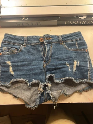Blå slitna jeansshorts med fransar - Säljer ett par blå jeansshorts med slitna detaljer och fransig kant. Shortsen har klassisk femficksmodell, bälteshällor och dragkedja med knapp. Perfekta för varma dagar och ger en avslappnad vibe.